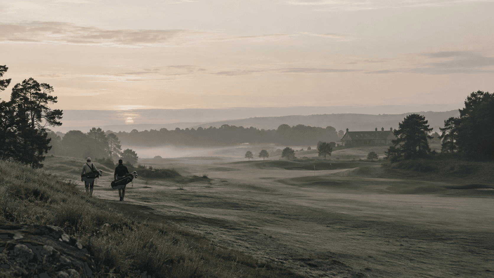 Two golfers carrying their bags walk a misty fairway at sunrise, the clubhouse in the distance.